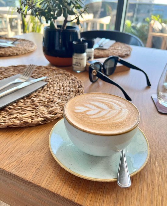 A cup of latte art featuring a leaf design sits on a wooden table next to black sunglasses and a woven placemat, with a potted plant in the background.