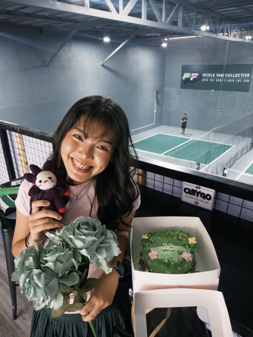 A smiling woman with long dark hair holds a knitted sheep plushie and a bouquet of sage green roses, standing next to a green birthday cake decorated with '23'.