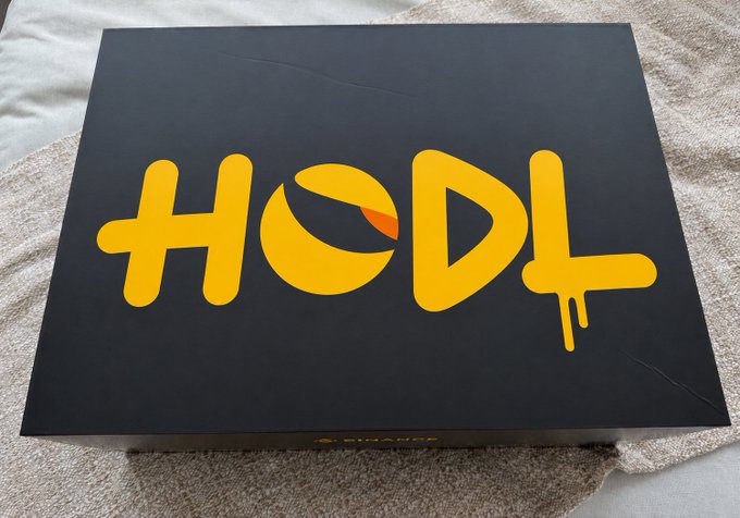 A black box or package rests on a tan towel, prominently displaying the word "HODL" in bold, dripping yellow graffiti-style letters, visually reinforcing the crypto slang term referenced in the post about the $LUNC token.