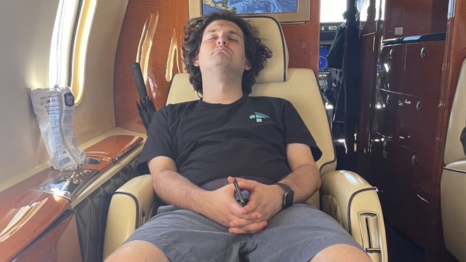 A man in a black t-shirt sleeps peacefully in a reclined leather seat inside a private jet, surrounded by wood-paneled interiors and luxury cabinetry. The image humorously illustrates the relaxed posture of someone unbothered by the financial turmoil described in the post about Alameda Research moving $15.2M in SOL to BitGo custody wallets following FTX's bankruptcy.