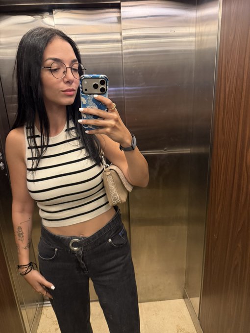A woman with long dark hair and round glasses takes a mirror selfie in an elevator, wearing a striped crop top and black jeans while holding a smartphone with a blue patterned case.