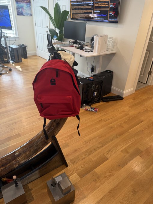 A red backpack sitting on a wooden chair in a home office space with hardwood floors, dual monitors displaying code/terminals on the wall, a desktop PC tower underneath the desk, and a potted plant nearby.