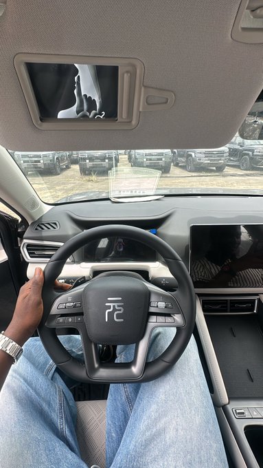 A person sits behind the wheel of a BYD vehicle, viewed from the driver's perspective showing the steering wheel with the Chinese character "元" (Yuan), the digital dashboard, and a rearview mirror display, with a row of parked cars visible through the windshield. The image captures a hands-on car shopping experience, embodying the post's message to "Go out, touch cars."