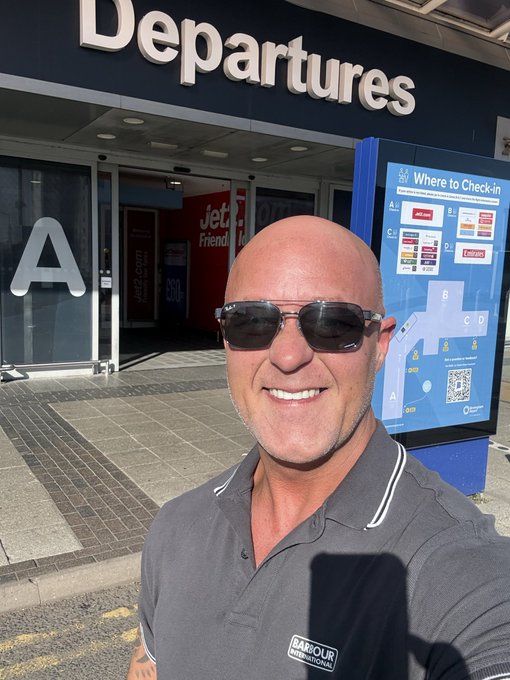 A bald man wearing sunglasses and a gray Barbour polo shirt takes a selfie outside an airport Departures terminal.