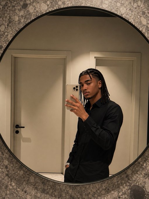 A young man with braided hair, dressed in a black shirt, takes a mirror selfie using a smartphone.