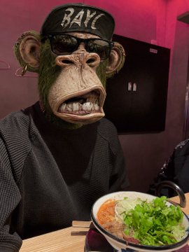 A monkey wearing sunglasses and a "RAVE" cap sits at a table with a steaming bowl of ramen topped with greens, set against a pink-lit background.