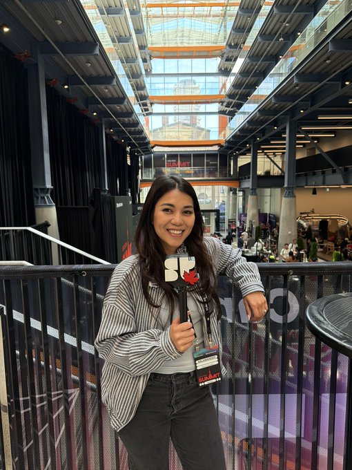 A smiling young woman with long dark hair wearing a striped shirt and SXSW badge lanyard, leaning on a railing in what appears to be a convention center atrium.