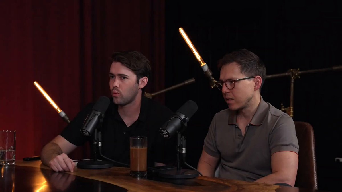 The image shows two men sitting at a table with microphones in front of them, appearing to be engaged in a podcast or radio show. The background features a red curtain, which adds color to the scene. There are also cups placed on the table, suggesting that they might be enjoying some beverages during their conversation.
