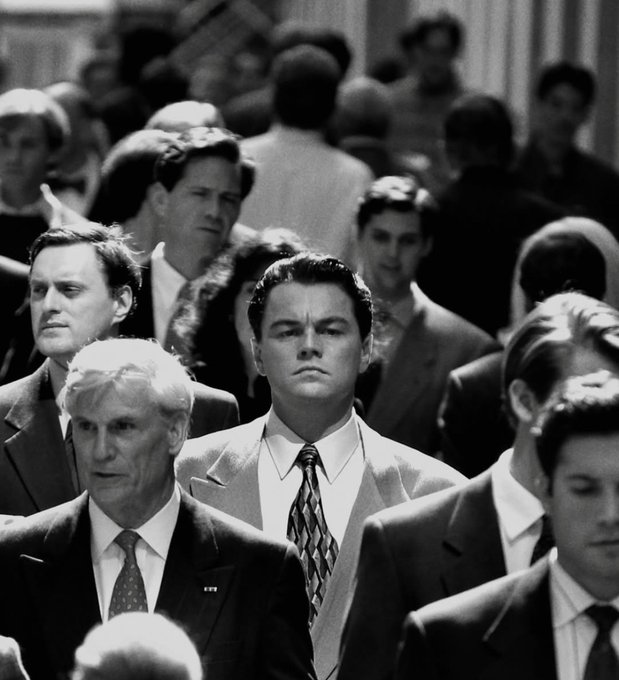 A black and white photo from the movie The Wolf of Wall Street showing Leonardo DiCaprio in a light suit and patterned tie, standing stoically in a crowd of businessmen.