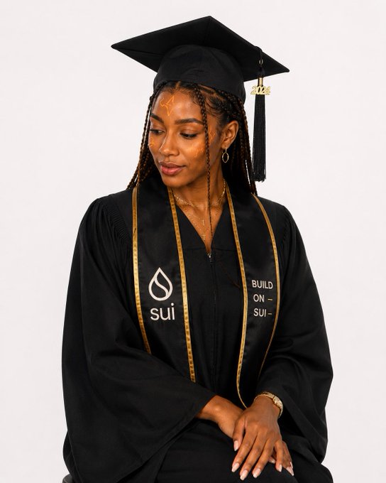 A young woman wearing a graduation cap and gown looks down with a serene expression, her black robe featuring the "sui" logo and the phrase "BUILD ON SUI" in gold lettering. The image playfully connects to the Sui Network post by using graduation attire to symbolize the successful completion of Perpetuals V2's security audit, marking the protocol's "graduation" to mainnet.