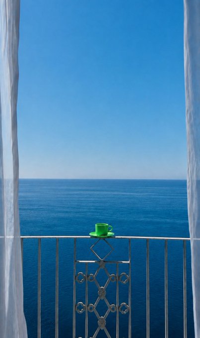 A bright green espresso cup rests on the railing of a seaside balcony, framed by flowing white curtains as calm blue ocean stretches endlessly under a clear sky. The serene view perfectly captures the tranquil morning ("GM") vibe referenced in the post, evoking the relaxed lifestyle that stable USDC earnings could help fund.