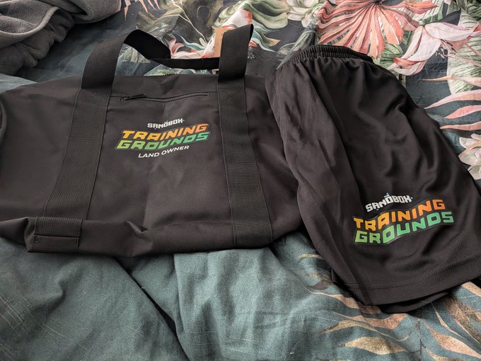 In the image, there are two items - a black bag with "Training Grounds" written on it and a pair of shorts. The bag appears to be made from canvas material and has a handle for carrying. The shorts are black and have green and yellow text on them that says "Training Grounds". The objects are placed next to each other, suggesting they might belong together or be related in some way.
