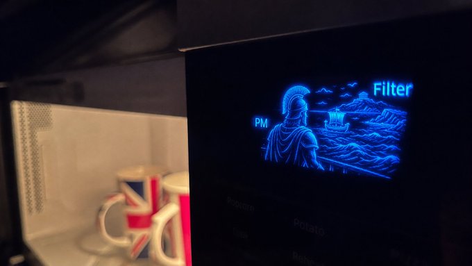 A digital screen displaying an illuminated blue illustration of a Roman soldier and a ship, labeled 'PM' and 'Filter', with coffee mugs visible on a shelf in the blurred foreground.