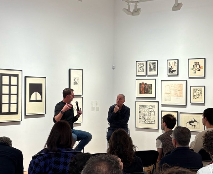 Two men sit on stools in an art gallery discussing with an audience, surrounded by framed artworks including black and white geometric prints on the left and a grid of mixed framed pieces on the right, as they introduce digital art and CryptoPunks to a new audience at ISLAA.