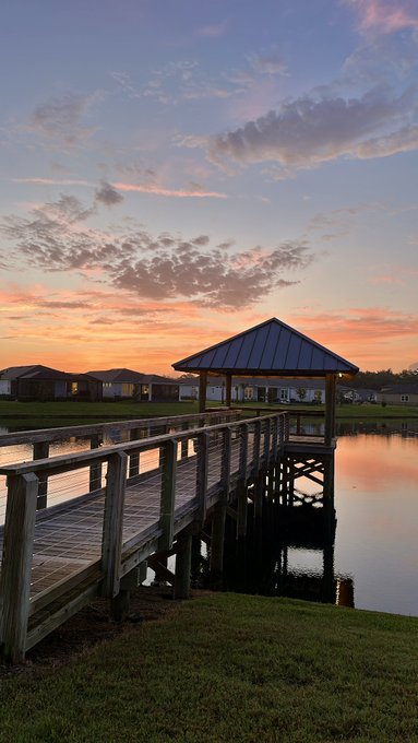 A scenic photograph of a wooden pier and gazebo extending over a calm lake at sunset, with houses visible in the background and pink and orange clouds in the sky.