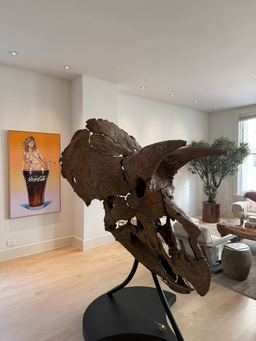 This image features a Triceratops skull, with prominent horns pointing upwards, humorously complementing the "horns up" sentiment of the post. A painting of a woman in a Coca-Cola glass is visible in the background.