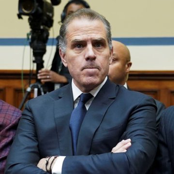 Hunter Biden, wearing a dark suit and blue tie, sits with his arms crossed during what appears to be a formal congressional hearing, with a camera operator visible in the background.
