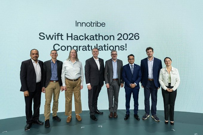 Eight people stand on stage in business attire at the Innotribe Swift Hackathon 2026 Congratulations ceremony, with one woman on the right proudly holding a glass trophy. The image captures the awards moment referenced in the post celebrating the hackathon winners, including @StanChart and @PictetGroup, alongside the runners-up.