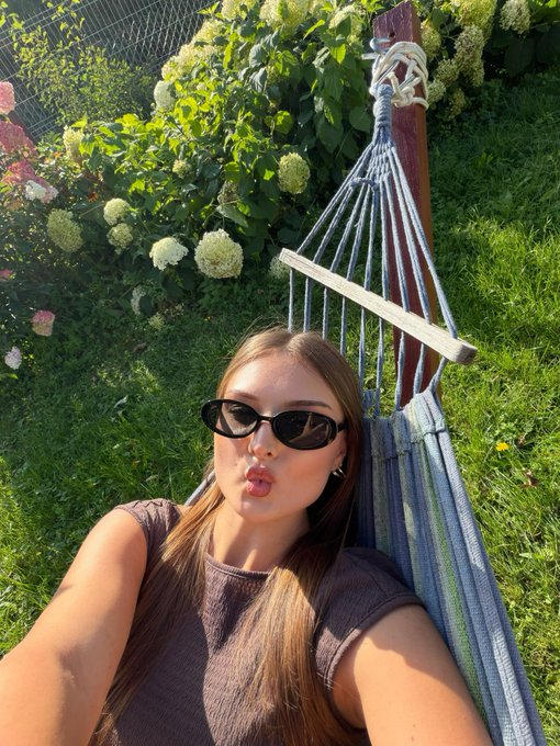A young woman with sunglasses poses for a selfie while lying in a striped hammock in a lush garden with white hydrangeas.