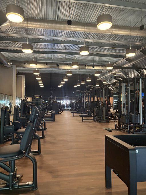 An empty, modern gym interior features rows of weight machines and exercise equipment under pendant lighting and an industrial-style ceiling.