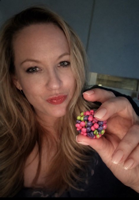 A selfie of a blonde woman holding up a cluster of colorful small berries (blueberries and pink/red berries) close to the camera.
