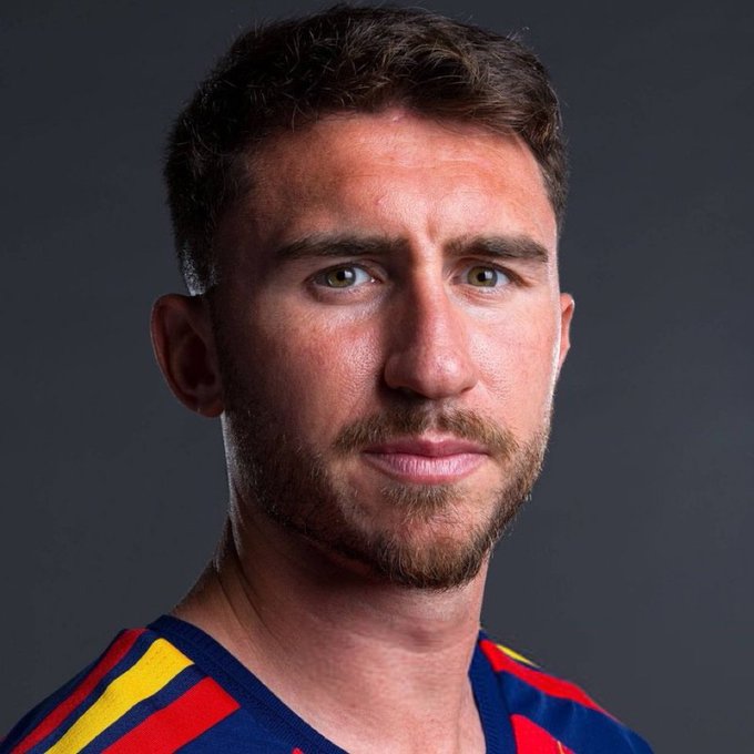 A close-up portrait of a man with short brown hair and a beard, wearing a blue shirt with red and yellow stripes, against a plain gray background.