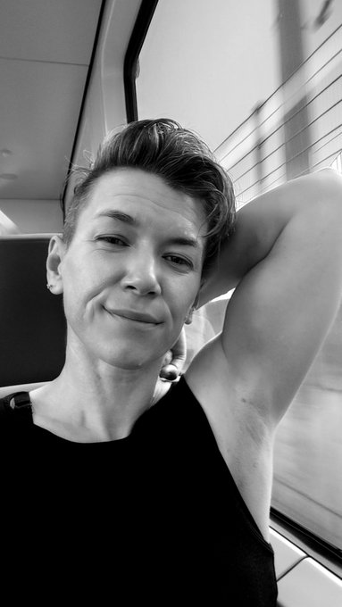 A black and white selfie of a person with short hair wearing a dark sleeveless top, resting their arm behind their head and looking directly at the camera.