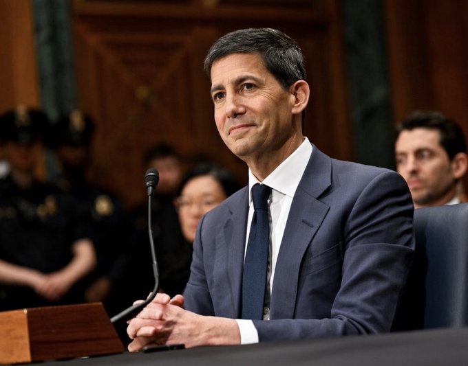 A photograph of a man in a dark suit seated at a hearing table with a microphone, appearing to be in a formal government or senate setting with uniformed personnel in the background.