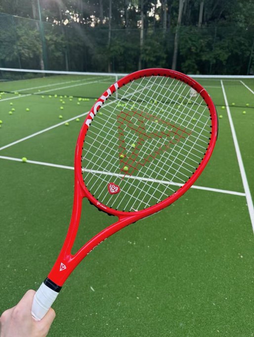 A red tennis racket held up against a green tennis court scattered with many yellow tennis balls.