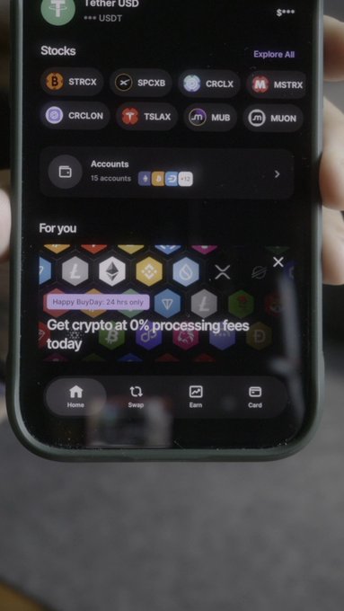 The image shows a smartphone displaying the Ledger Wallet app home screen, featuring a "Happy BuyDay: 24 hrs only" banner promoting 0% processing fees for buying digital assets, along with various stock tickers like STRCX, SPCXB, CRCLX, and MSTRX visible in the Stocks section. The promotional offer highlights zero processing fees for the next 24 hours when purchasing crypto through supported providers like MoonPay, Coinify, and others as mentioned in the post.