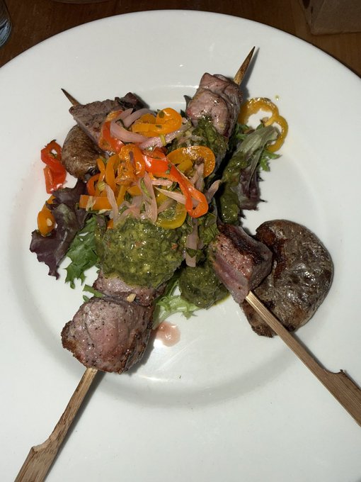 A white plate holds two wooden skewers of grilled steak and sausage over a bed of mixed greens, topped with a green herb sauce and sliced red onions, orange peppers, and yellow peppers.