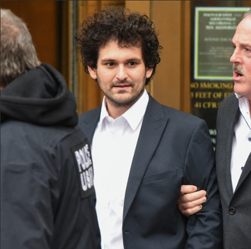A man with curly hair wearing a dark suit and white shirt stands flanked by individuals, including a person in a black jacket labeled "PRESS" with "CSD" visible, appearing to be escorted outside a courthouse with "NO SMOKING" signs posted on the wall. The image relates to the caption showing Sam Bankman-Fried as he pursues legal action at the US Supreme Court to overturn his conviction and 25-year sentence for an $8 billion fraud case.