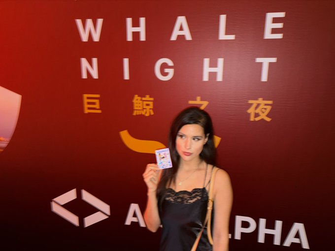A woman in a black satin top holds up a card while posing in front of a red wall displaying "WHALE NIGHT" in English and "巨鲸之夜" in Chinese, with an "ALPHA" logo visible below—fitting the "chinesemaxxing trencher" Bitcoin Conference Asia post about attending a crypto whale event in Asia.