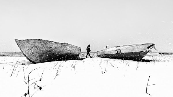 A black and white photograph of a person standing between two large, weathered wooden boats stranded on a snowy, barren landscape.