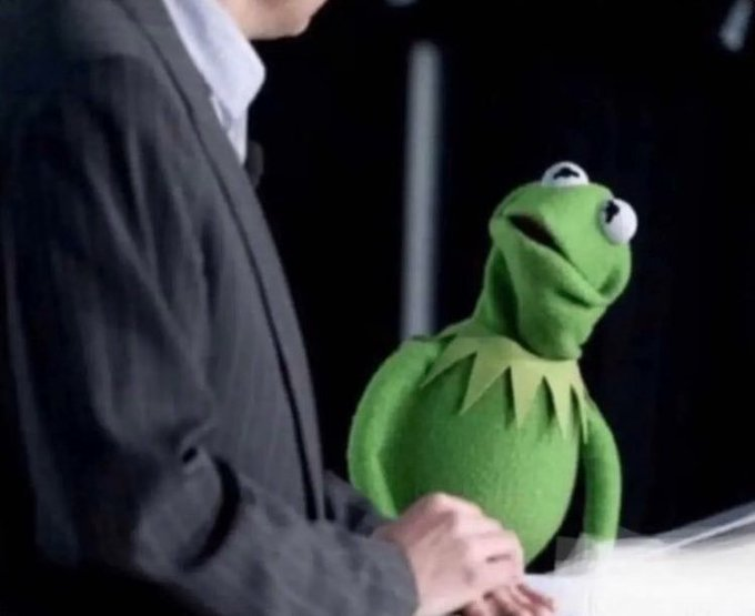 Kermit the Frog sits next to a person wearing a dark suit, appearing to be at a desk or table.
