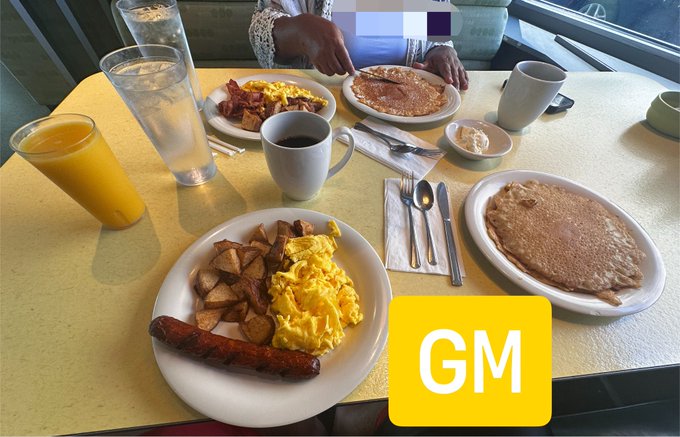 A breakfast spread on a yellow table at what appears to be a diner, featuring plates with eggs, bacon, sausage, hash browns, and pancakes, along with orange juice, coffee, and water, with a yellow 'GM' (Good Morning) sticker overlay.