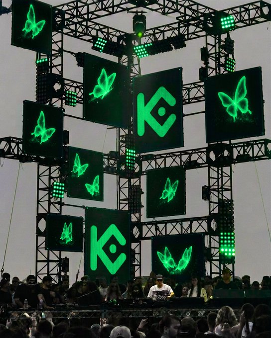 A concert stage features large green-lit screens displaying the "KC" logo and glowing butterflies, towering above the DJ booth and crowd. The bold visual branding reinforces the post's confident message—that Koze (KC) commands the biggest stages without needing an introduction.
