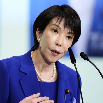 A close-up photograph of Japanese politician Sanae Takaichi wearing a blue blazer and pearl necklace while speaking at a podium with microphones.