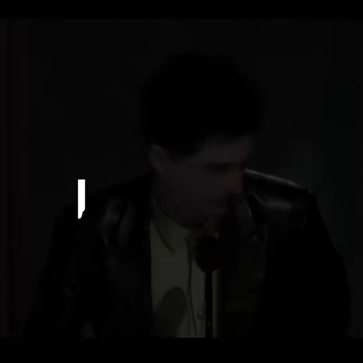 A dark, low-lit image shows a person in a leather jacket speaking into a microphone, with a small white shape visible on the left side.