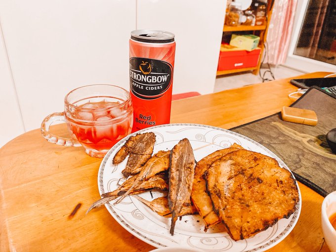 A meal of fried meat and what appears to be fried fish or chicken is served on a plate next to a glass mug of iced drink and a can of "Strongbow Red Berries" apple cider on a wooden table.
