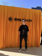 A person wearing sunglasses and black clothing poses in front of an orange BNB Chain branded backdrop.