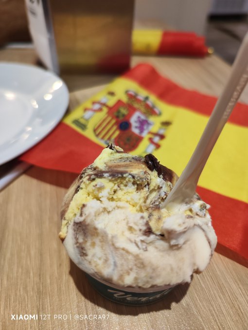 A cup of gelato or ice cream topped with cake pieces sits on a wooden table with a Spanish flag visible in the background; watermark reads 'XIAOMI 12T PRO | @SACRA97'.