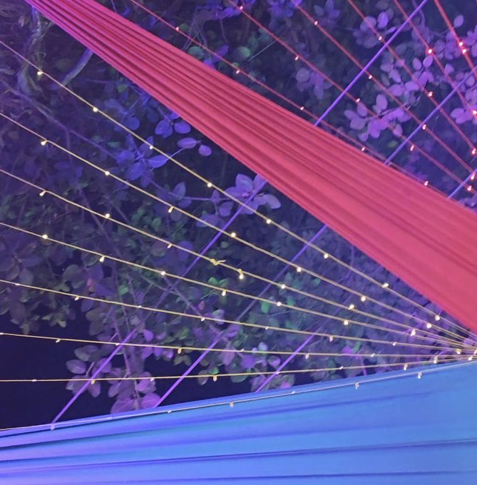 The image features a large blue and purple tent with strings of lights strung across its surface. The tent appears to be set up for an event or gathering, possibly a wedding or outdoor party. The strings of lights are arranged in various patterns and directions, creating a visually appealing display that adds to the festive atmosphere.