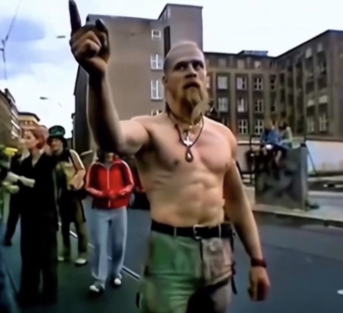 A shirtless, muscular animated character with a goatee holds up a gun pointed skyward on a city street, while bystanders watch. The caption "I said higher" humorously suggests the character is commanding someone to raise their hands or weapon even higher, fitting the defiant, larger-than-life pose.