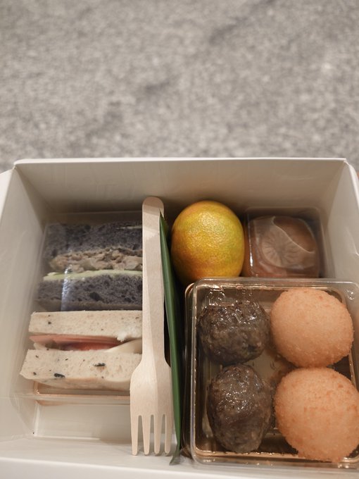 An open bento box contains sandwiches, an orange, and assorted round snacks including meat balls and rice balls, with a wooden fork included.