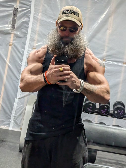 A muscular, bearded man wearing a "UNAFFECTED" cap takes a mirror selfie in a gym, flexing his heavily muscled arms while checking his phone, embodying the post's message about earning results through hard work—described as "TRT Peptide free." Dumbbells and gym equipment are visible in the background, reinforcing the dedication to natural, effort-based fitness progress.