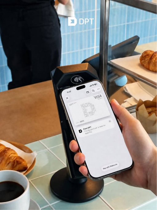 A hand holding an iPhone to make a contactless Visa payment via Apple Pay at a payment terminal branded 'DPT,' with a coffee cup, croissant, and bakery items visible in the café setting.