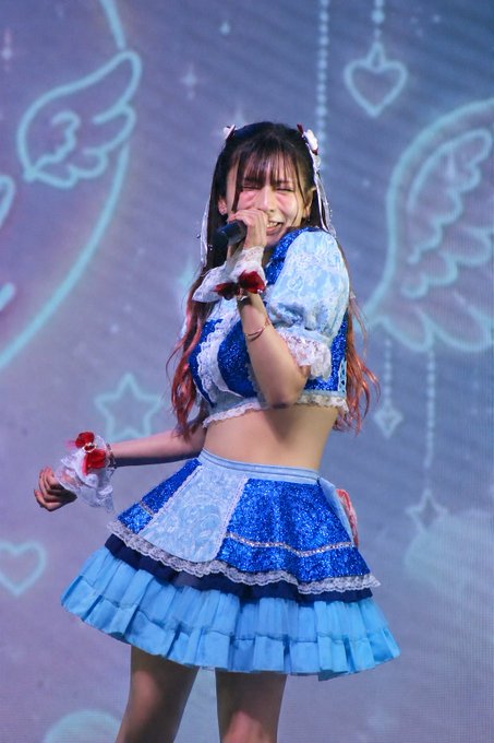 A young performer in a blue and white frilly outfit with red hair accessories sings into a microphone on stage.
