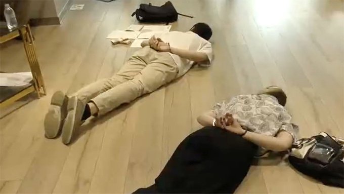 Two people lie face down on a wooden floor; a person in light clothing and another in a patterned shirt rest near scattered papers and bags.