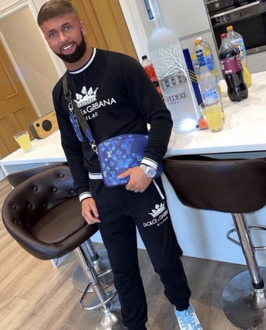 A bearded man with an edited cartoon smile stands in a kitchen wearing a black Dolce & Gabbana sweatshirt and matching pants, holding a blue Louis Vuitton crossbody bag, with a Belvedere vodka bottle on the counter behind him.